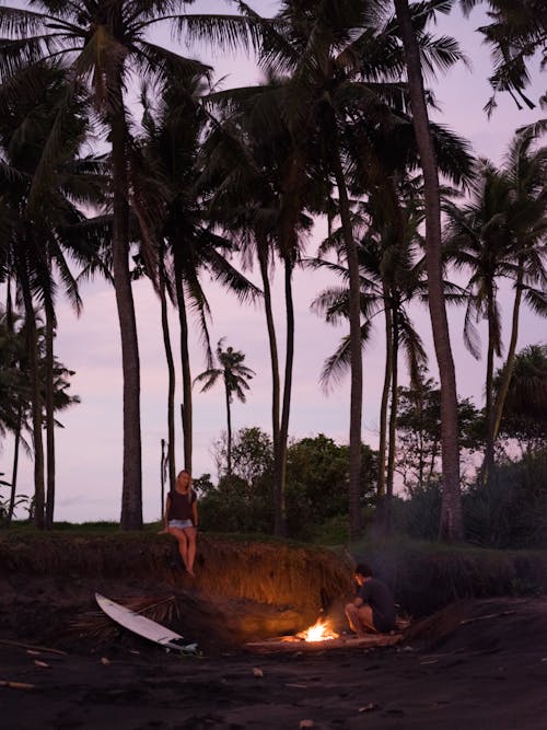 Unrecognizable young travelers relaxing near campfire while spending time together on beach among palm trees on tropical resort