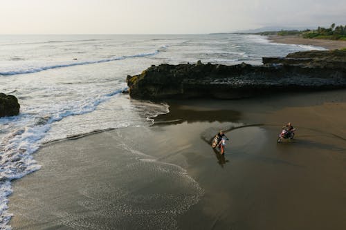 Remote view of anonymous people driving motorbikes on wet sand of coastline with rocks during active holidays in sunset