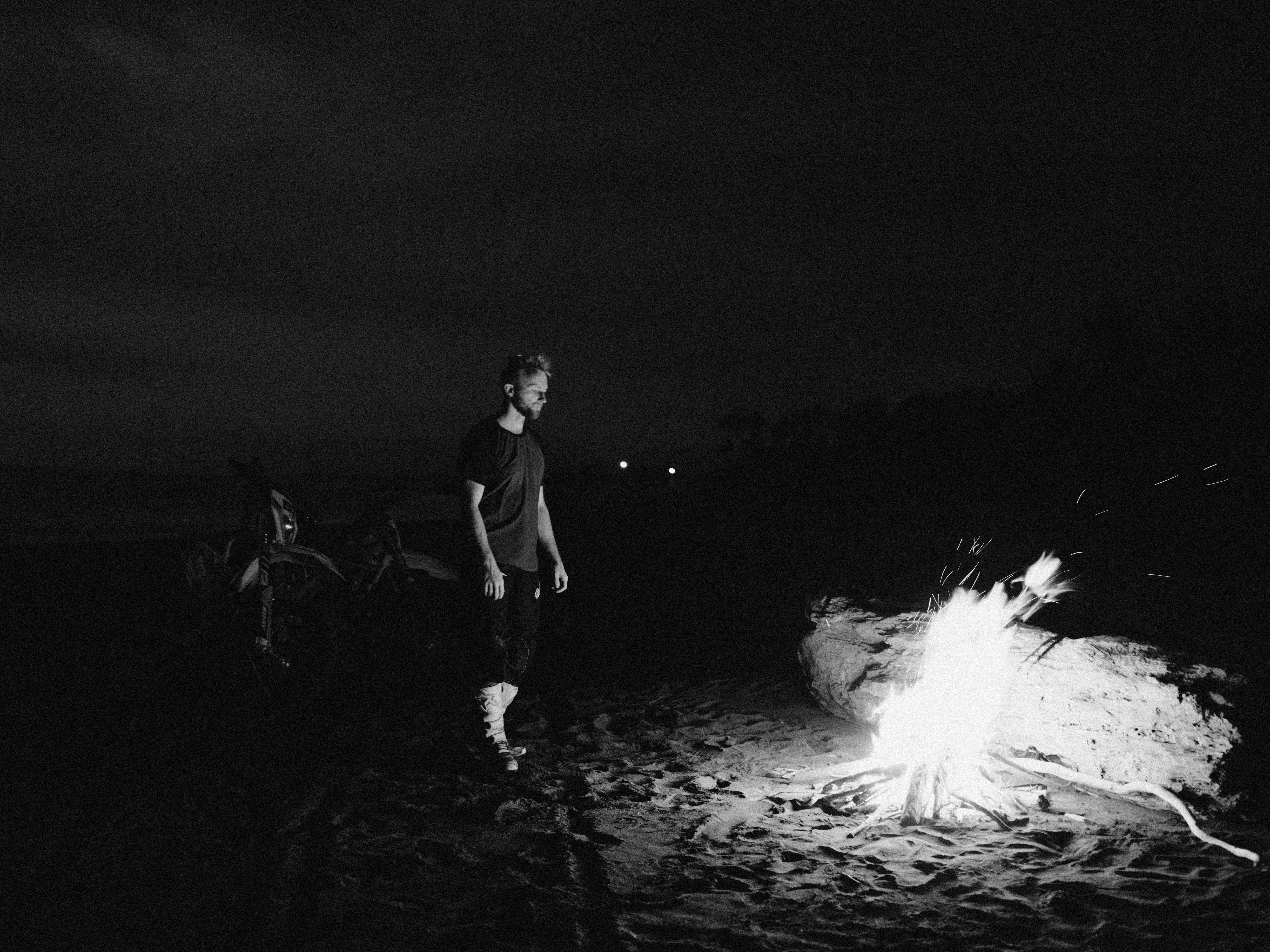 夜晚，一名孤独的男子站在海滩的篝火旁，身旁停着一辆摩托车