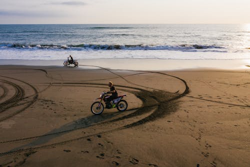 Distant anonymous bikers driving motorcycles on wet sandy beach with tracks in sunset time during active travel