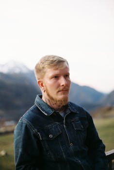 A contemplative man with a beard poses in a denim jacket against a mountain backdrop.