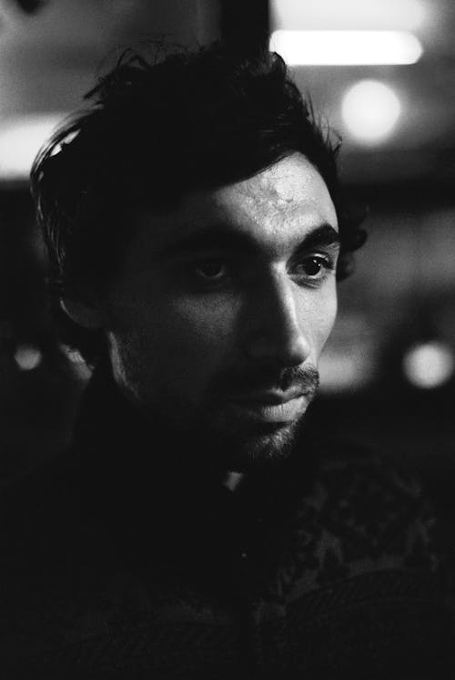 Black and white portrait of serious bearded guy with stylish hairdo on dark blurred background with night lights looking away thoughtfully