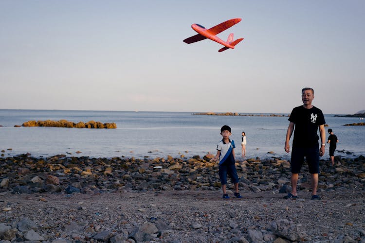 Ethnic Father And Son Launching Toy Plane On Beach At Sunset