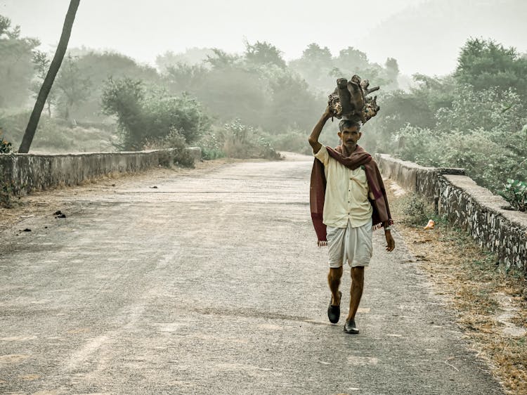 Serious Ethnic Man Walking Along Path In Village With Heavy Items On Head