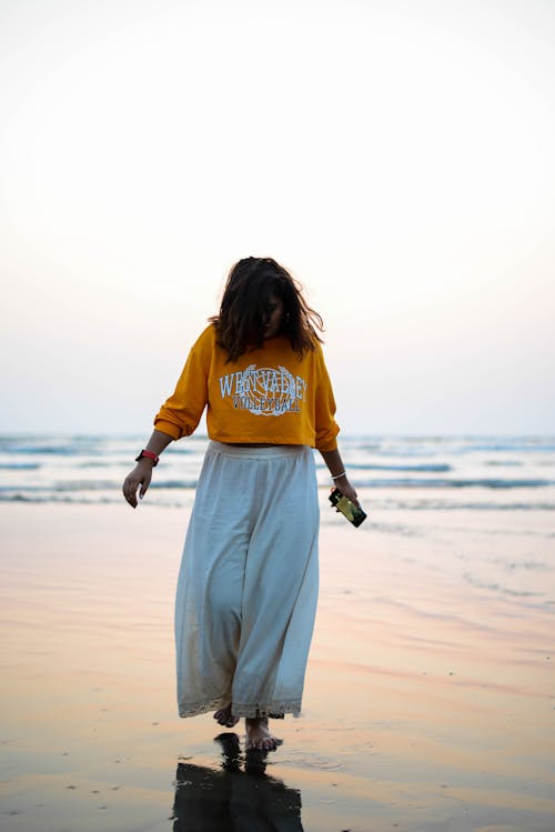 Woman in Orange Long Sleeve Shirt and White Skirt Walking on Beach