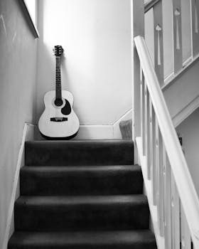 Monochrome image of an acoustic guitar placed on indoor carpeted stairs, featuring classic design.