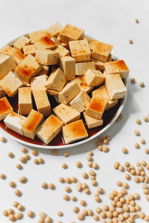 Free Appetizing tofu cubes drizzled with soy sauce on a white plate, surrounded by raw soybeans. Stock Photo