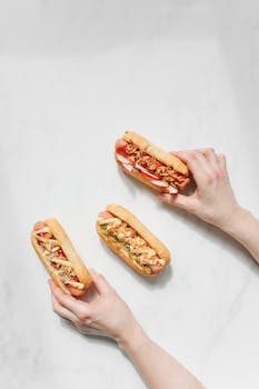 Overhead view of three gourmet hot dogs with toppings held by hands on a marble surface.