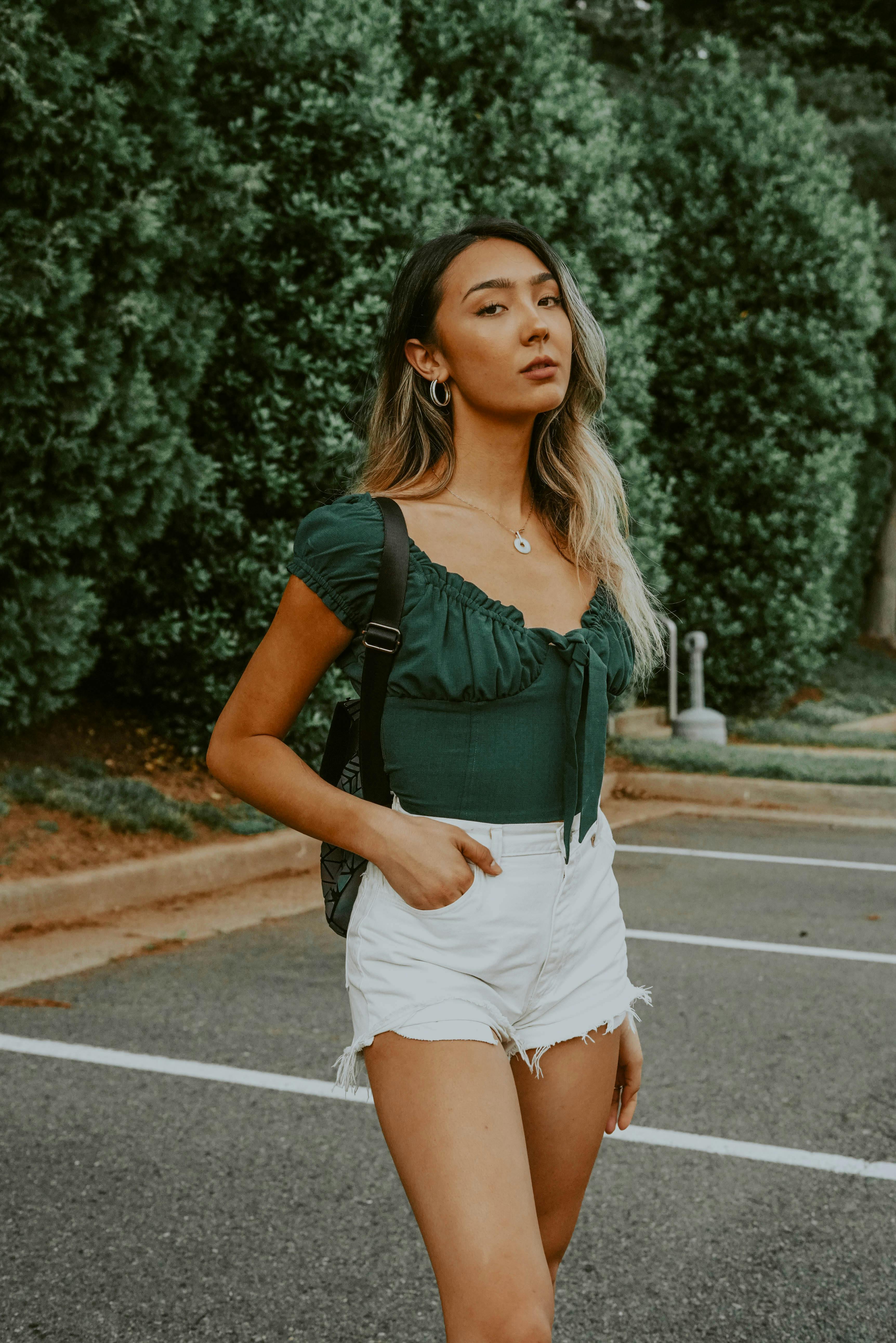 Serious Asian female with backpack standing on asphalt parking lot against green trees