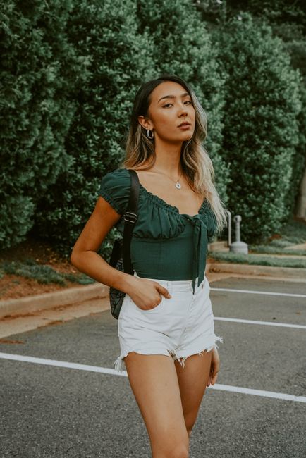 Serious Asian female with backpack standing on asphalt parking lot against green trees