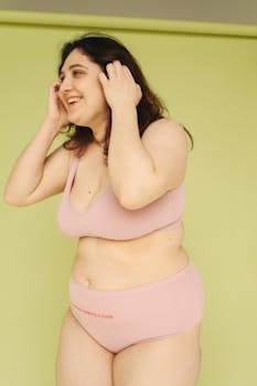 Smiling woman in pink lingerie embracing body positivity on green backdrop.