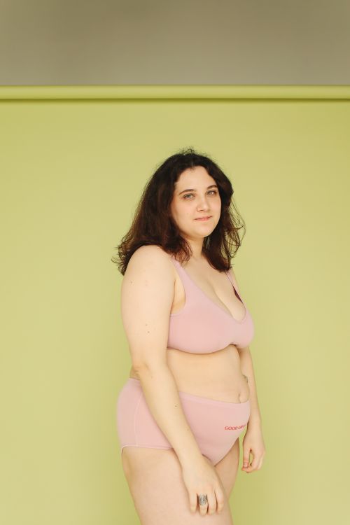Free Portrait of a confident woman wearing pink lingerie, standing against a light green backdrop, embracing body positivity. Stock Photo
