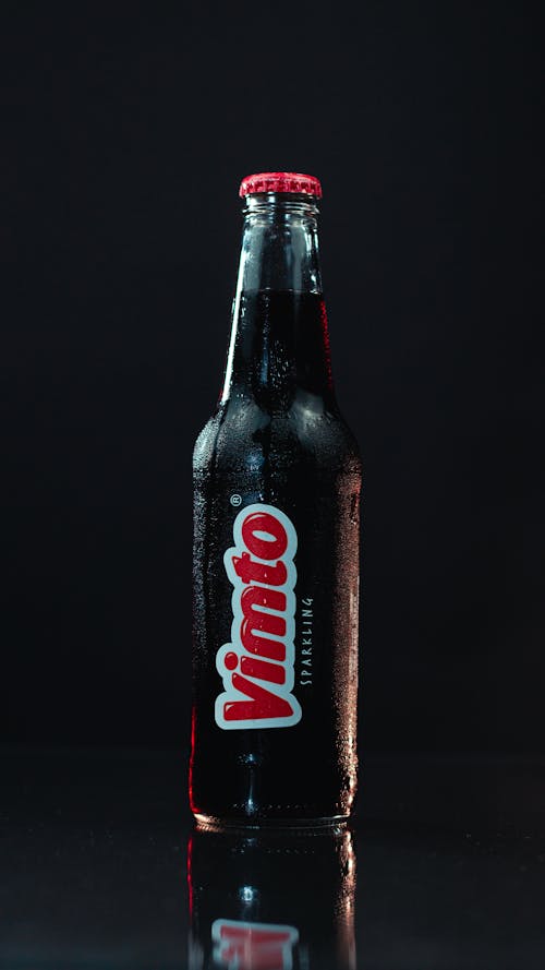 Glass transparent bottle of cold soft drink placed on black reflecting surface in dark room