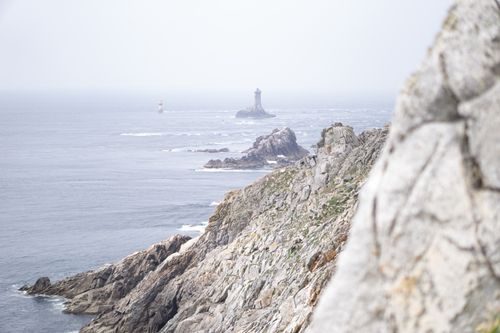 Free A tranquil seascape featuring rocky cliffs and a distant lighthouse on a foggy day. Stock Photo