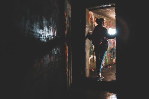 Free Back view of anonymous female with backpack standing in doorway of dark room near shabby painted wall of abandoned building Stock Photo