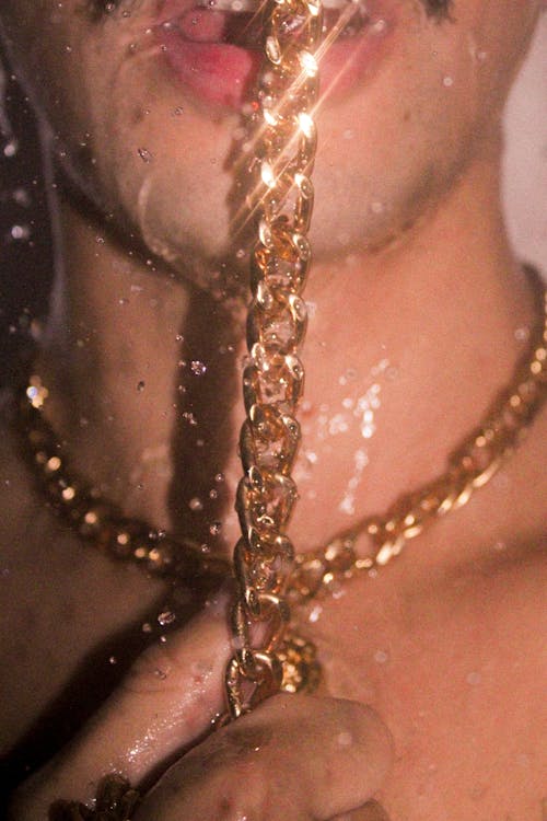 Faceless male with mustache standing under shower with shiny golden chain on neck and touching chain with lips while showering