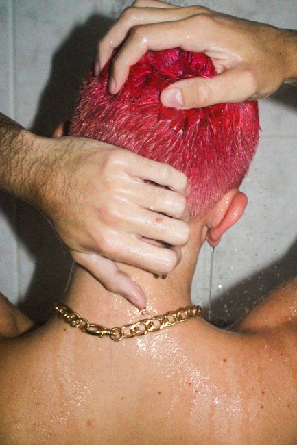 Back view of anonymous male with dyed hair standing under pouring water with hands on head and gold chain on neck
