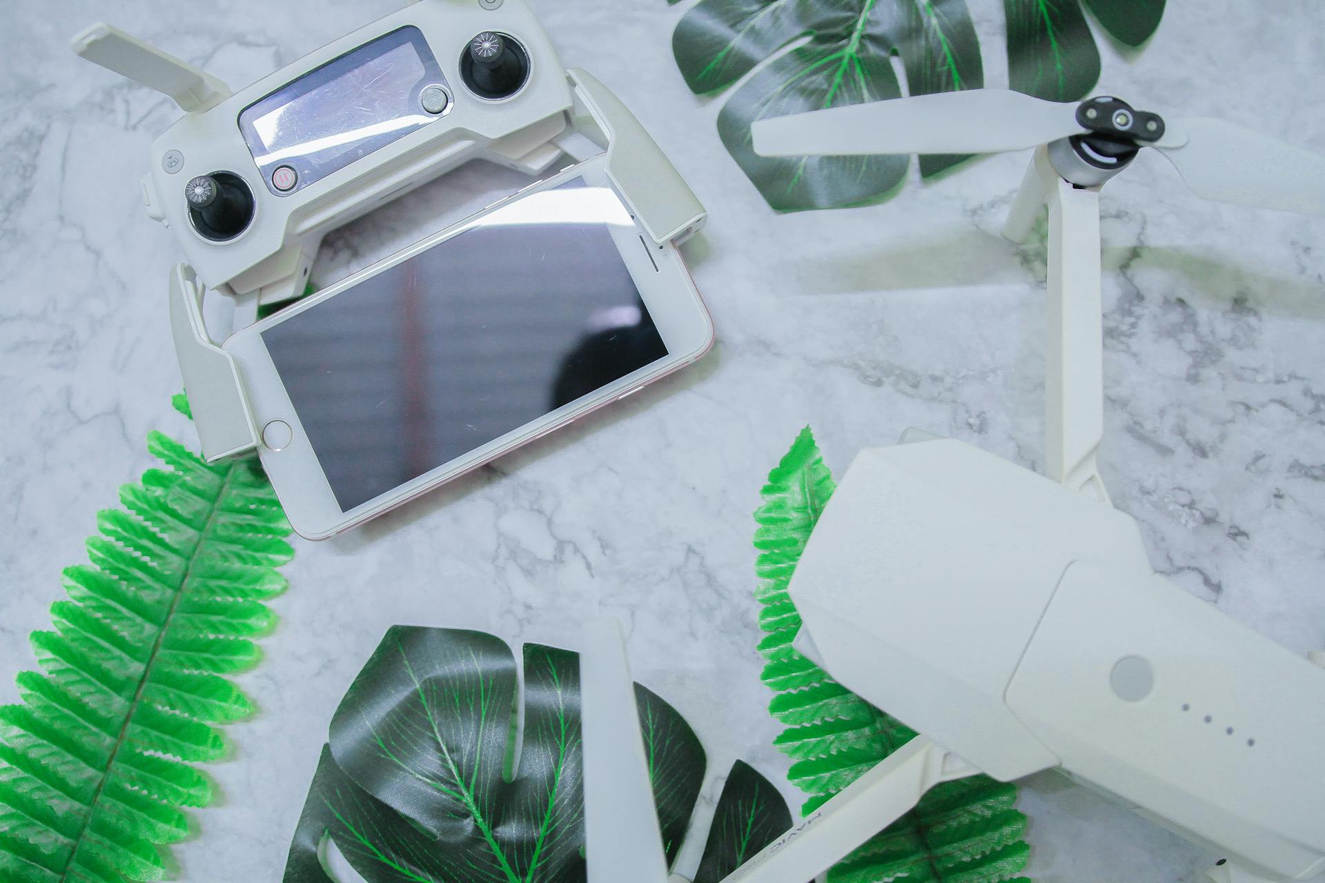 Flat lay of a drone controller and smartphone on marble with green leaves. High-tech setup.