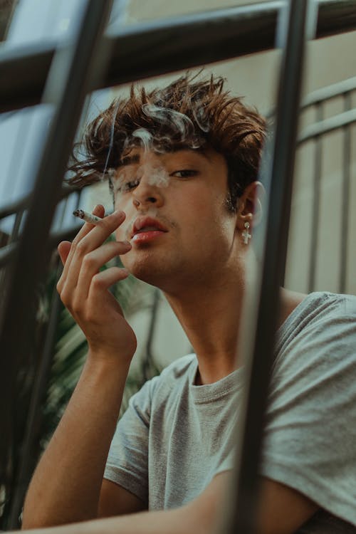Serious male in casual clothes sitting on stairs and smoking cigarette while looking at camera during summer day in city