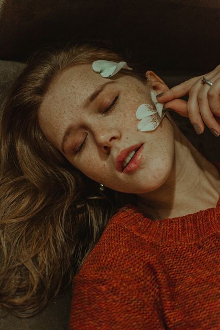 Dreamy female with blond hair in red sweater lying with closed eyes with petals of white flower on face during rest