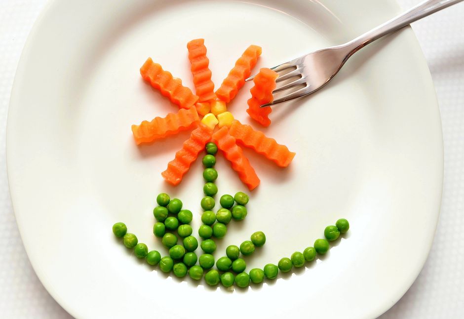 5 Maneiras de Usar a Criatividade na Alimentação Saudável 2 A playful flower made from carrots and peas on a white plate with a fork, showcasing creativity in food art.