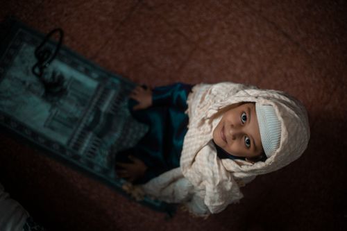 Free A young girl in a hijab smiling while sitting on a prayer mat, viewed from above, indoors. Stock Photo