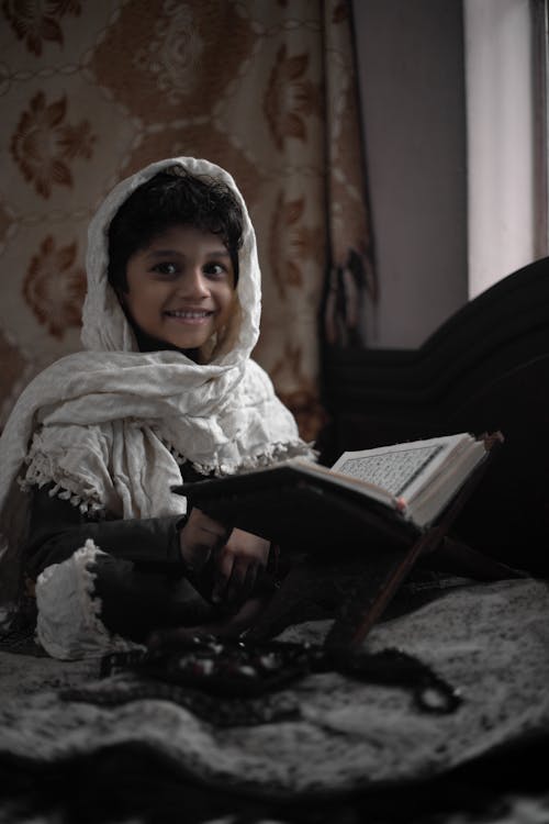 Little ethic child in white blanket sitting on bed in room and reading interesting book while looking at camera during daytime