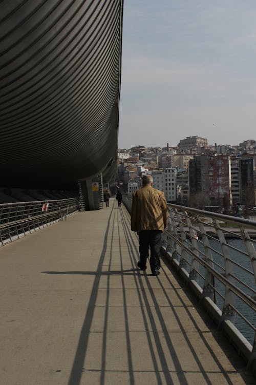 Back view of anonymous man in worn clothes walking across bridge over river in big city at sunny day