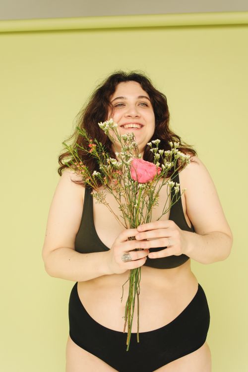 Free Smiling woman in underwear holding a flower bouquet against a pastel background, embracing body positivity. Stock Photo