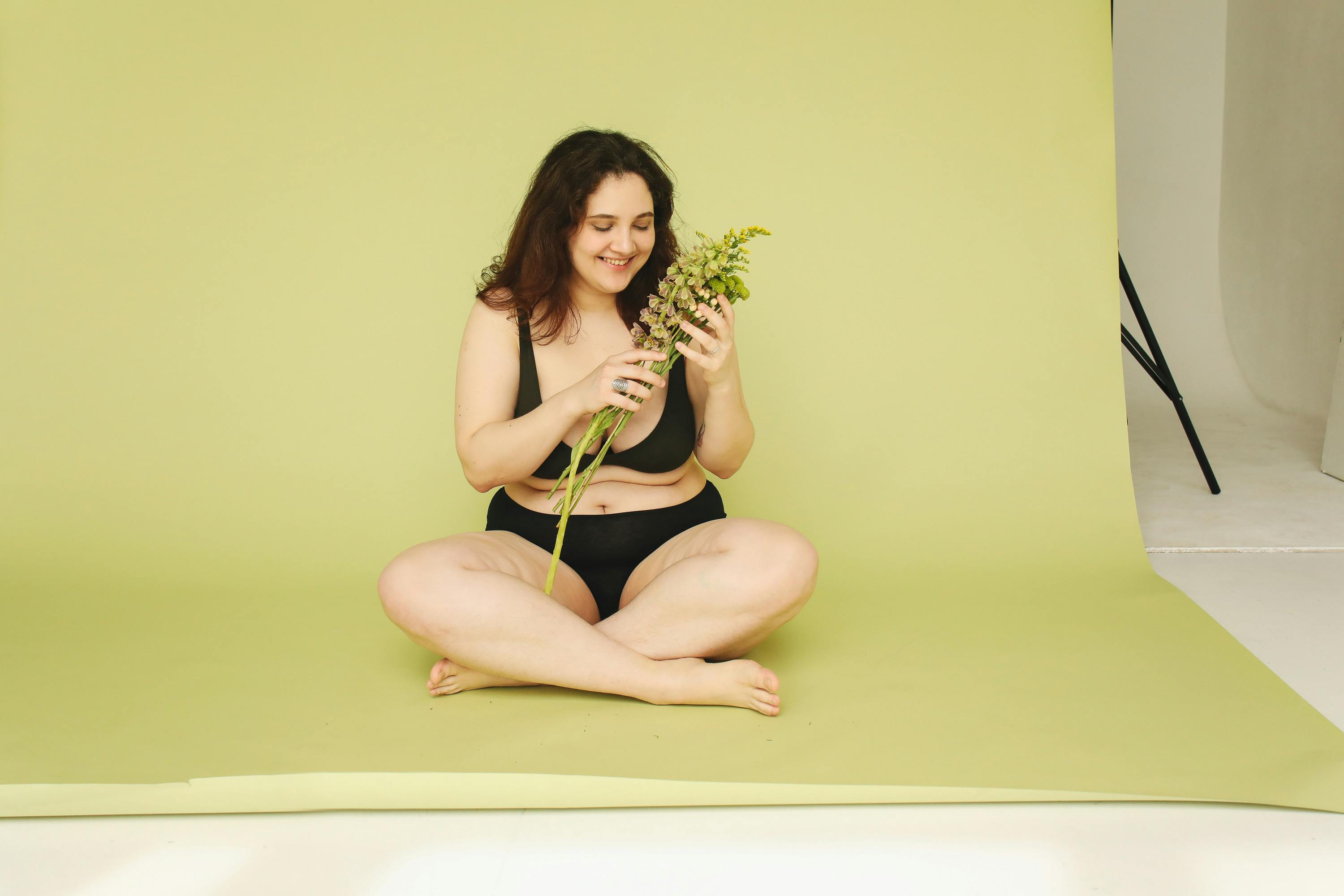 woman wearing black undergarments sitting on the yellow floor while looking at the stem of green plant she is holding