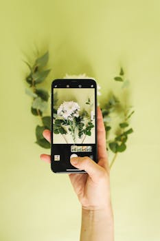 Hand holding a smartphone capturing a photo of flowers and leaves against a green backdrop.