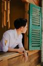 Side view of dreamy ethnic male in stylish apparel looking out of window of wooden house near decorative wind bell