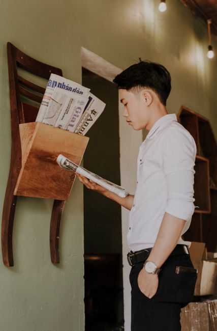 Side view of young trendy ethnic guy with wristwatch and hand in pocket reading newspaper in house Side view of young trendy ethnic guy with wristwatch and hand in pocket reading newspaper in house
