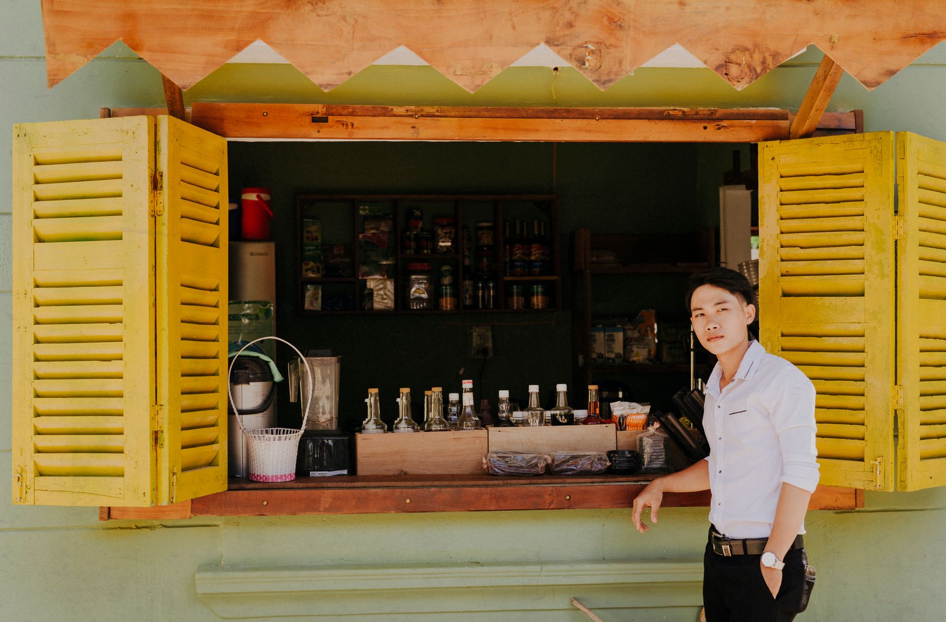 Man standing at an outdoor beverage stand with open yellow shutters offering a variety of drinks.