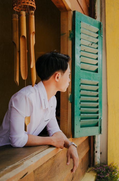 Side view of anonymous young contemplative Asian guy in elegant shirt looking away from wooden window