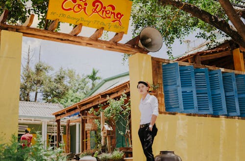 Low angle of wistful Asian guy in stylish clothes standing with hands in pockets near colorful wall of old house while looking away