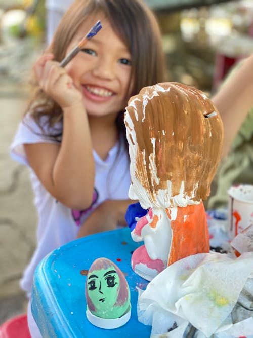 Free From above of crop smiling Asian girl with paint brush looking at camera while sitting at table with colorful painted doll and decorative egg Stock Photo
