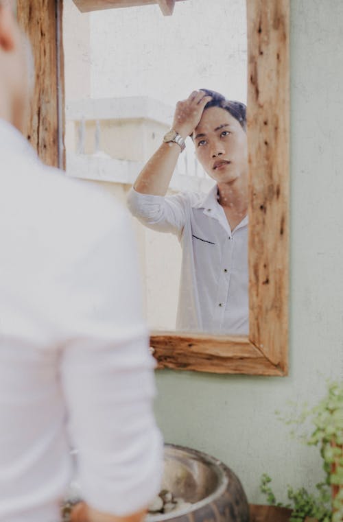 Back view of crop stylish pondering ethnic male touching hair while reflecting in mirror in house and looking away