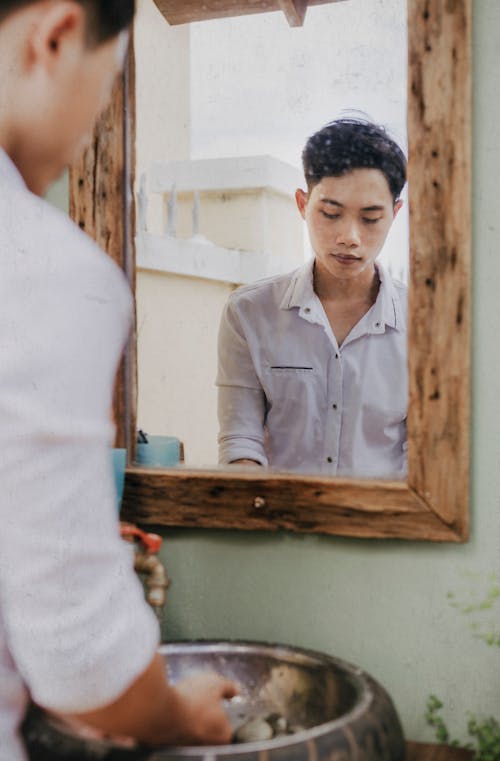 Back view of crop wistful Asian male in trendy wear washing hands in washstand while reflecting in mirror at home and looking down