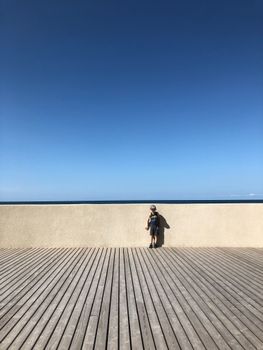 Anonymous little child standing on embankment near fence and ocean under bright sky