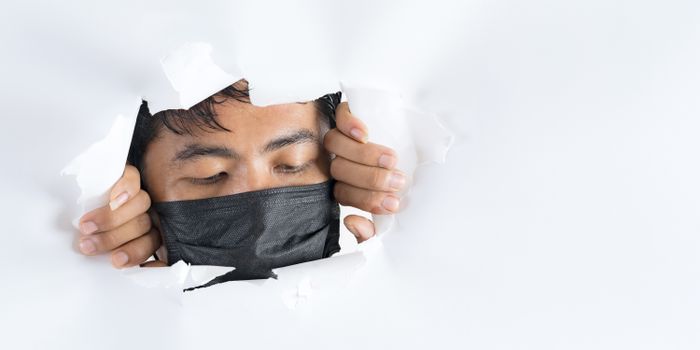 Crop anonymous ethnic male in face mask looking down behind ragged paper wall during COVID 19 pandemic