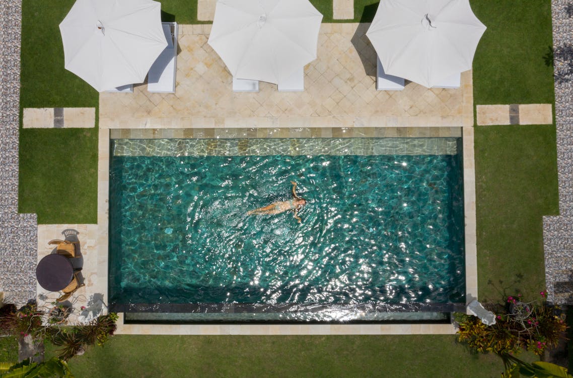 Drone view of anonymous female tourist relaxing in pool water near terrace with white parasols among green lawns in comfortable hotel zone during vacation