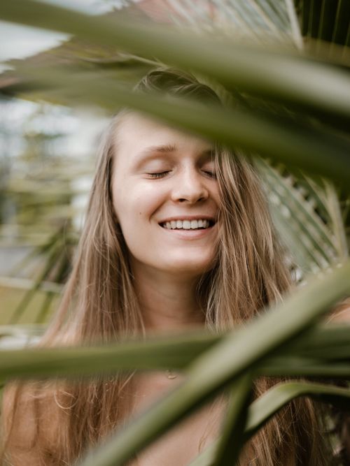 Free Smiling young woman with long blond hair closing eyes and laughing while standing near green plants in tropical garden Stock Photo