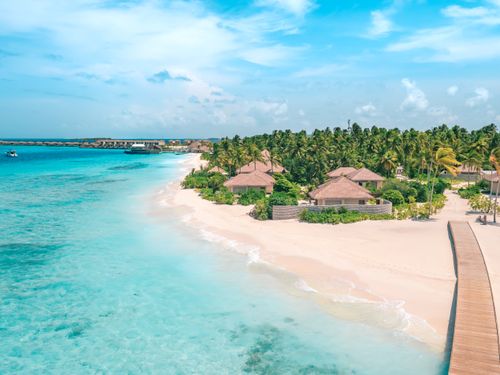 Free From above of pure ocean with sandy shore near wooden boardwalk and trees with small houses under blue cloudy sky Stock Photo