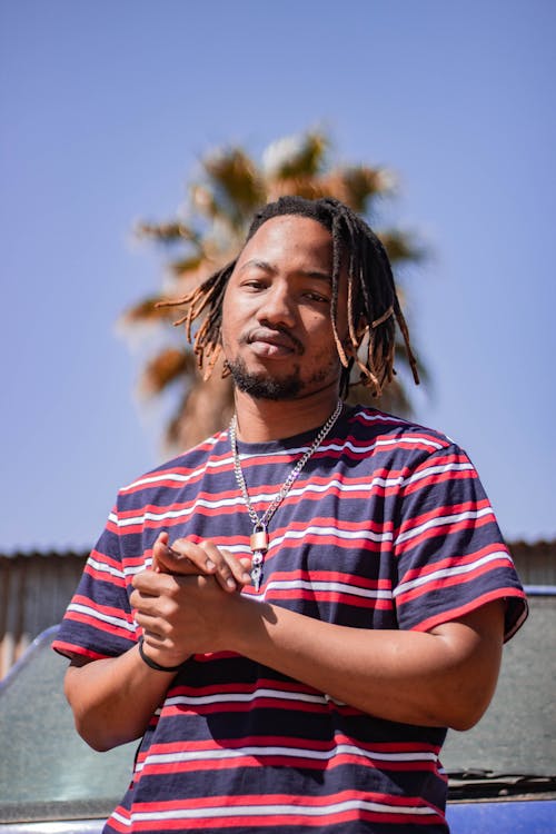 Low angle of stylish black male in striped t shirt and decorative pendant with dreadlocks looking at camera with clasped hands in daytime