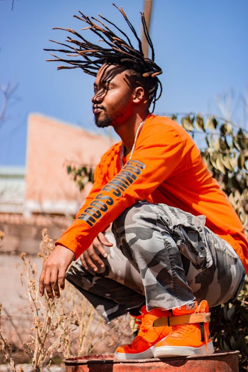 From below side view of trendy African American male with creative hairstyle in bright footwear squatting with closed eyes under blue sky