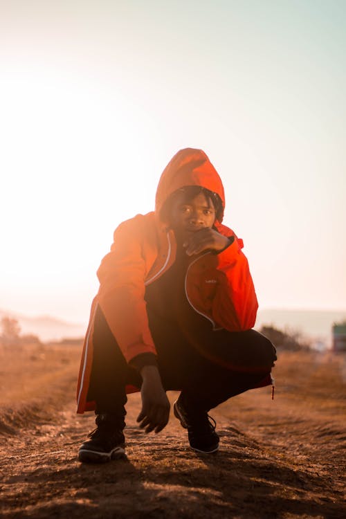 Ground level of stylish African American guy in hood touching face while squatting on dry terrain and looking away under sky at sundown