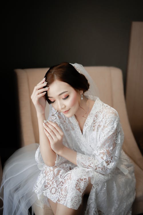 Free From above of reflective ethnic woman with makeup in bridal dress touching hair while sitting in chair with closed eyes Stock Photo