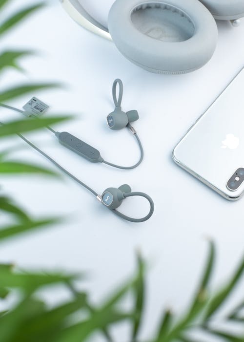 From above through green plant leaves arrangement of modern wired earphones and headphones placed on white desk near silver mobile phone