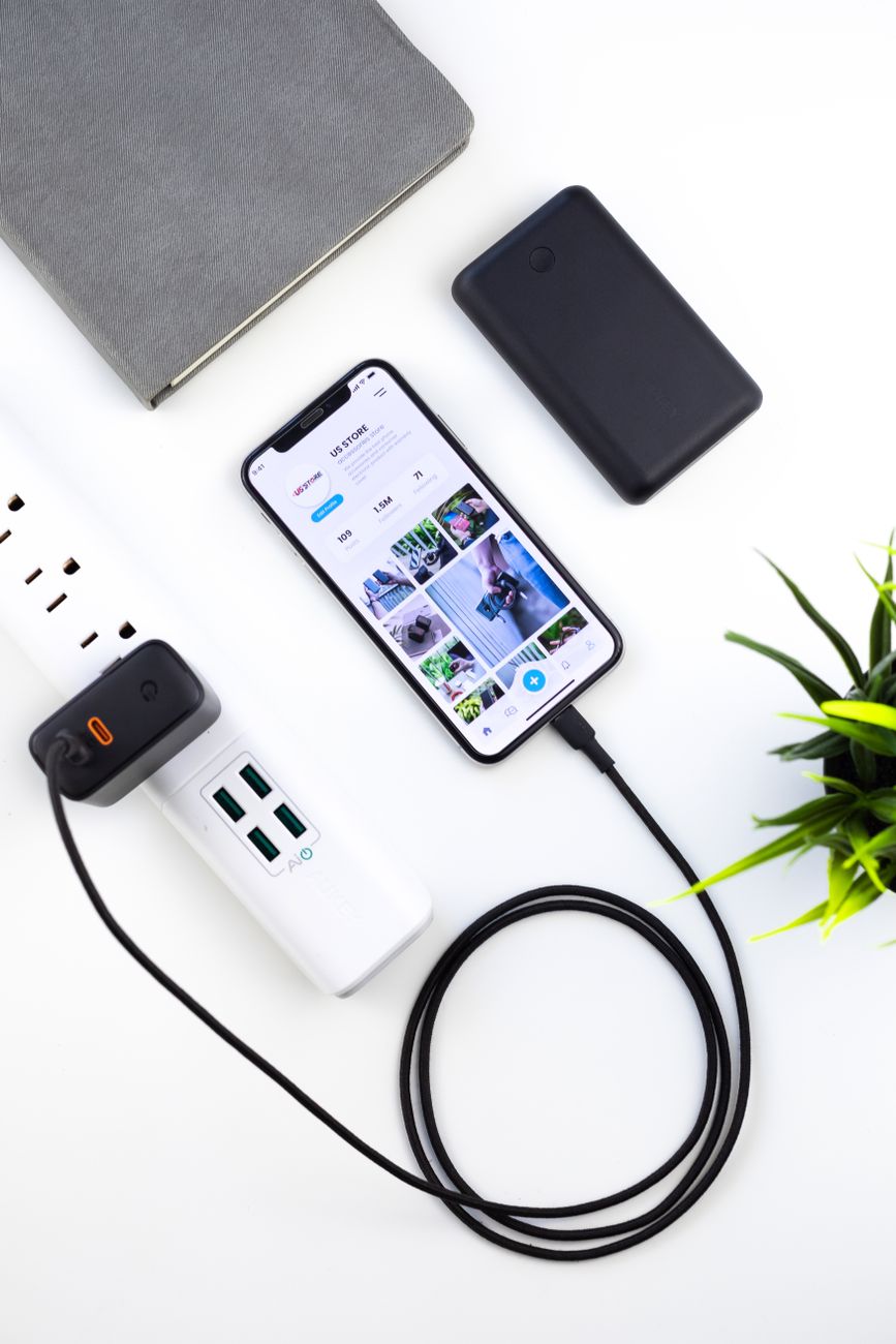 Top view composition of modern switched on mobile phone recharging on white table near tablet and green plant